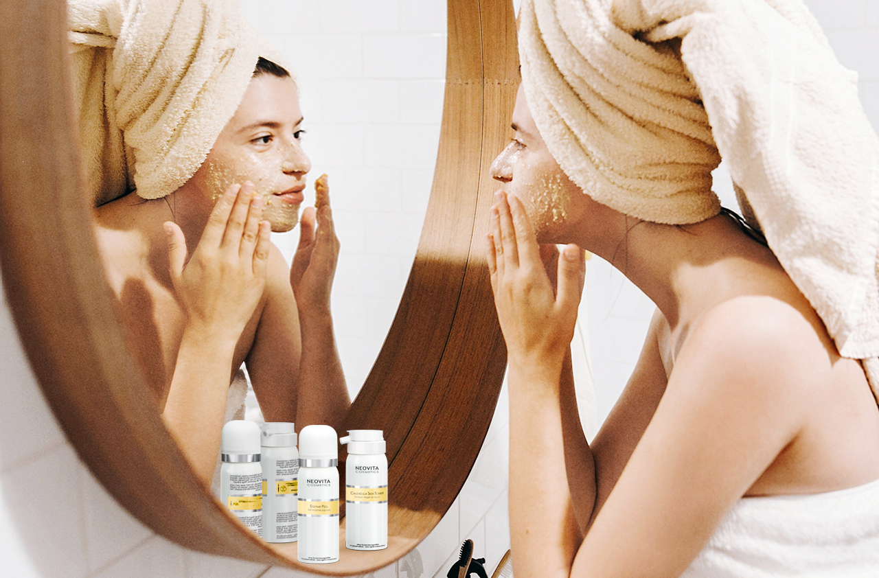 Frau mit Handtuch auf dem Kopf trägt im Badezimmer sanft ein Peeling von NEOVITA Cosmetics auf ihr Gesicht auf, Produkte der Enzyme Peel Serie im Vordergrund.